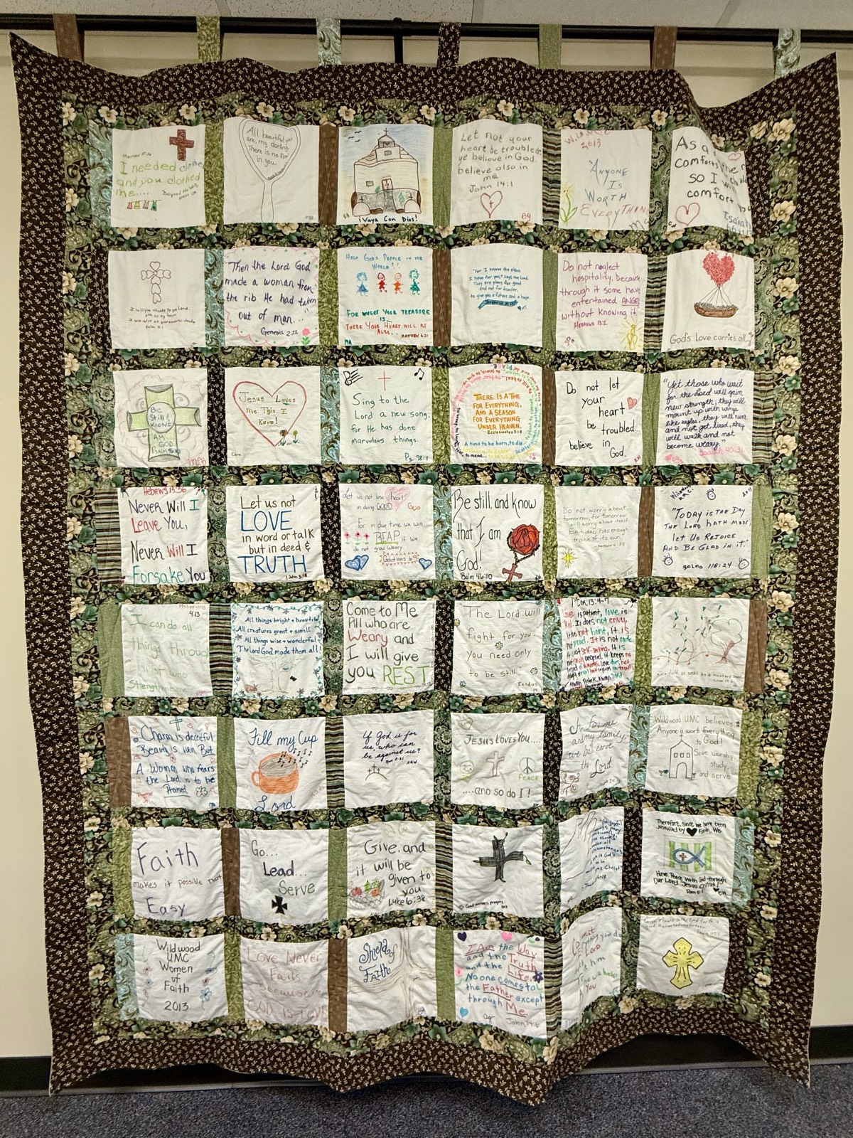 A large quilt hanging on a classroom wall, made of forty-eight cream-colored patches in eight rows, each hand-lettered with a Bible verse or phrase and some illustrated with crosses, a church, a heart-shaped balloon and a teacup. Green and brown floral sashing separates the patches, framed by a dark brown floral border.